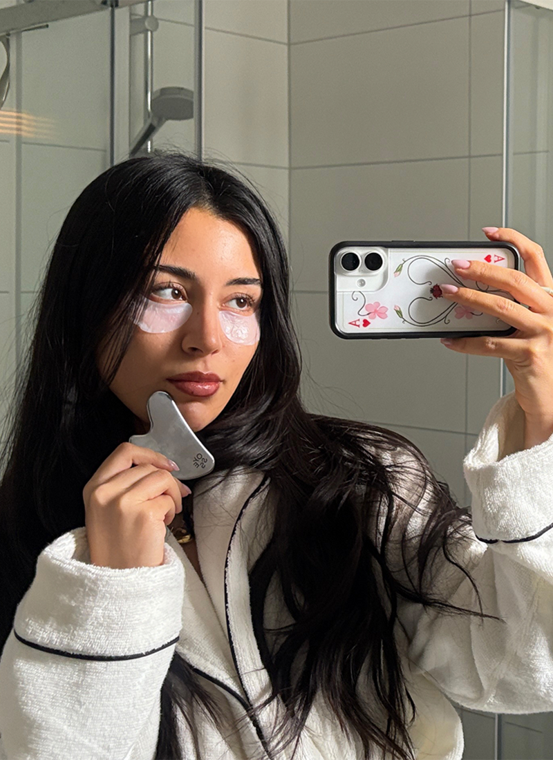 junge Frau die in einem Badezimmer steht, Eye Patches unter den Augen hat und einen Gua Sha in der einen Hand und das Telefon in der anderen Hand 