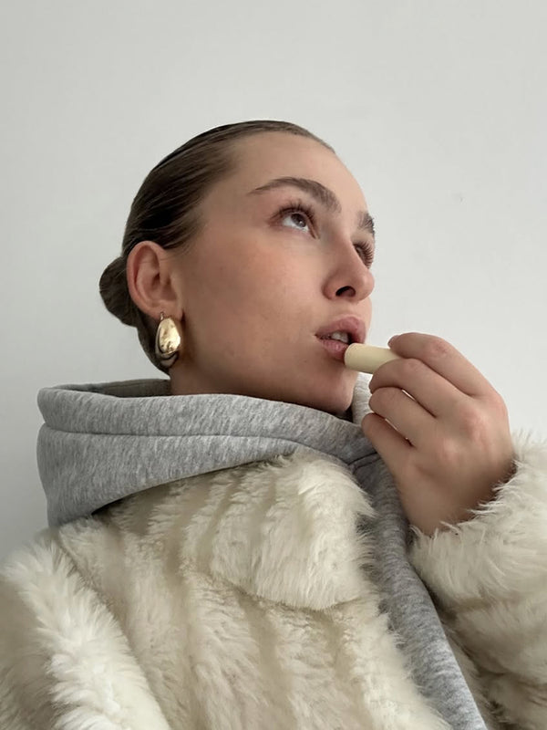 Portrait einer jungen Frau in cremefabenem Mantel mit einem cremefarbenen Lippenpflegestift in der Hand