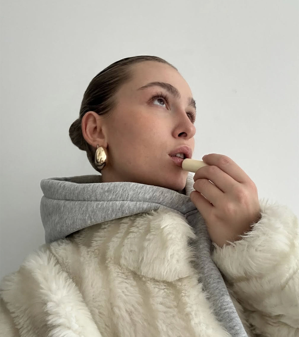 Portrait einer jungen Frau in cremefabenem Mantel mit einem cremefarbenen Lippenpflegestift in der Hand