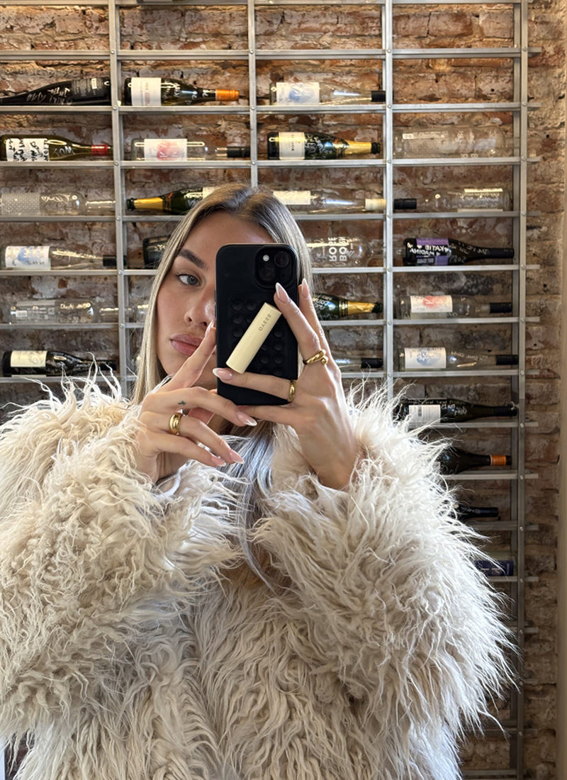 Foto einer jungen Frau in einem cremefarbenen Plüschmantel vor einer gepflasteren Wand mit einem Regal voller Flaschen die ein Telefon und ein einen cremefarbenen Lippenpflegestift in den Händen hält und ein Selfie macht 